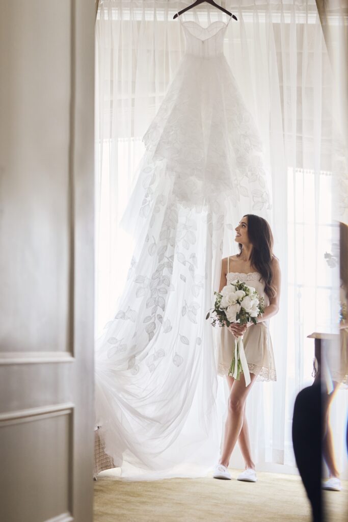 A woman standing with her wedding bouquet in front of her wedding gown, the moments before she is to be a bride